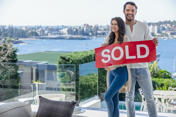 A young foreign couple stand inside their newly-bought Australian home with a sold sign